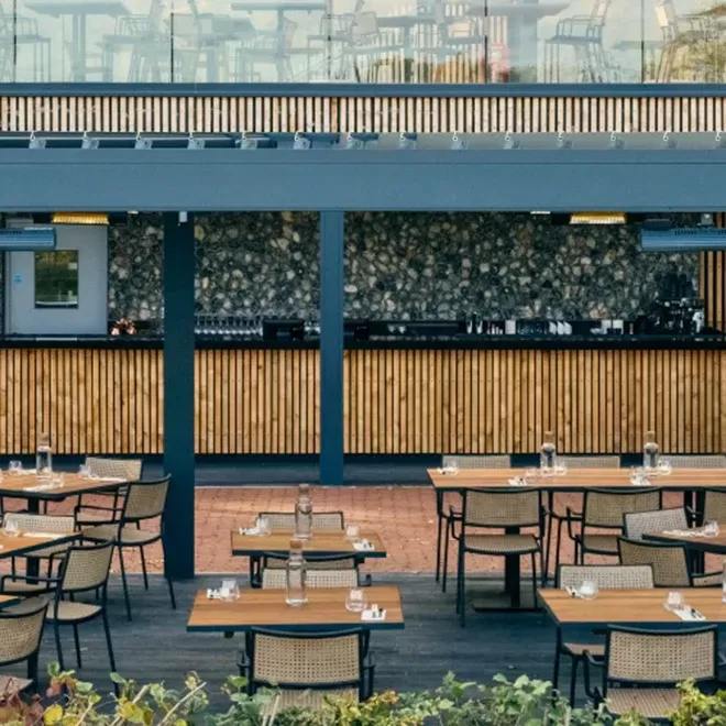 An outdoor restaurant features several wooden tables with chairs, each set with glasses and napkins. A bar with a stone facade and wooden slats is in the background, under a pergola. Lush greenery is visible in the foreground at The Rows & Vine at Ridgeview.