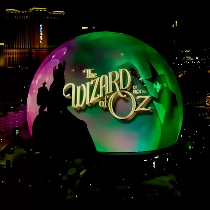 A large spherical building is illuminated at night with colorful lights and an image promoting The Wizard of Oz, showing silhouettes of characters and the title text against a vibrant backdrop.