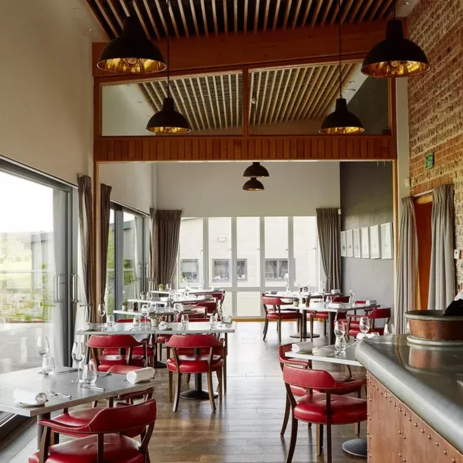 A modern restaurant interior with red cushioned chairs, neatly set tables, and large windows. The ceiling features wooden panels with hanging lamps, and an elegant bar is visible on the right. Natural light fills the spacious room at The Tasting Room at Rathfinnny.