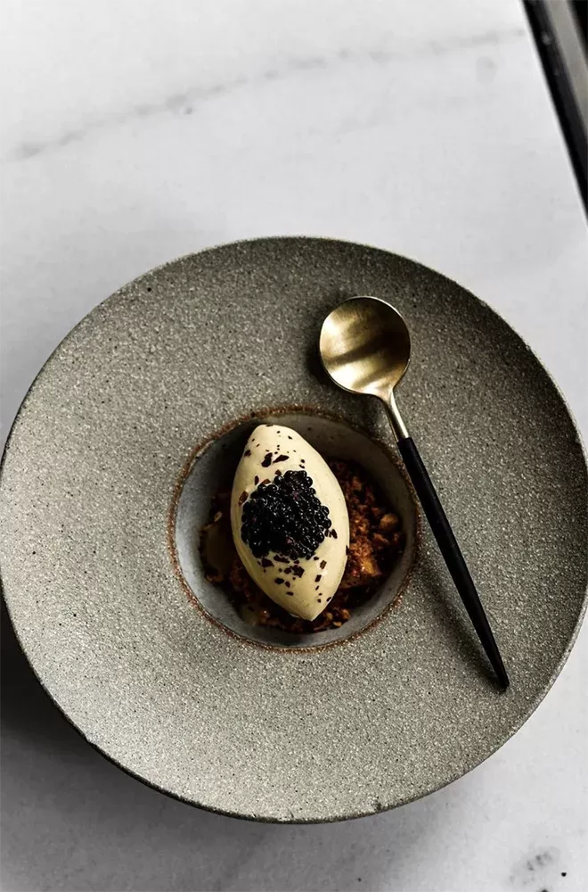 A dessert with a creamy scooped dollop topped with black caviar, presented on a bed of crispy crumbs in a gray, textured bowl. An elegant spoon with a golden bowl and dark handle rests on the bowls rim at The Water House Project, Bethnal Green.