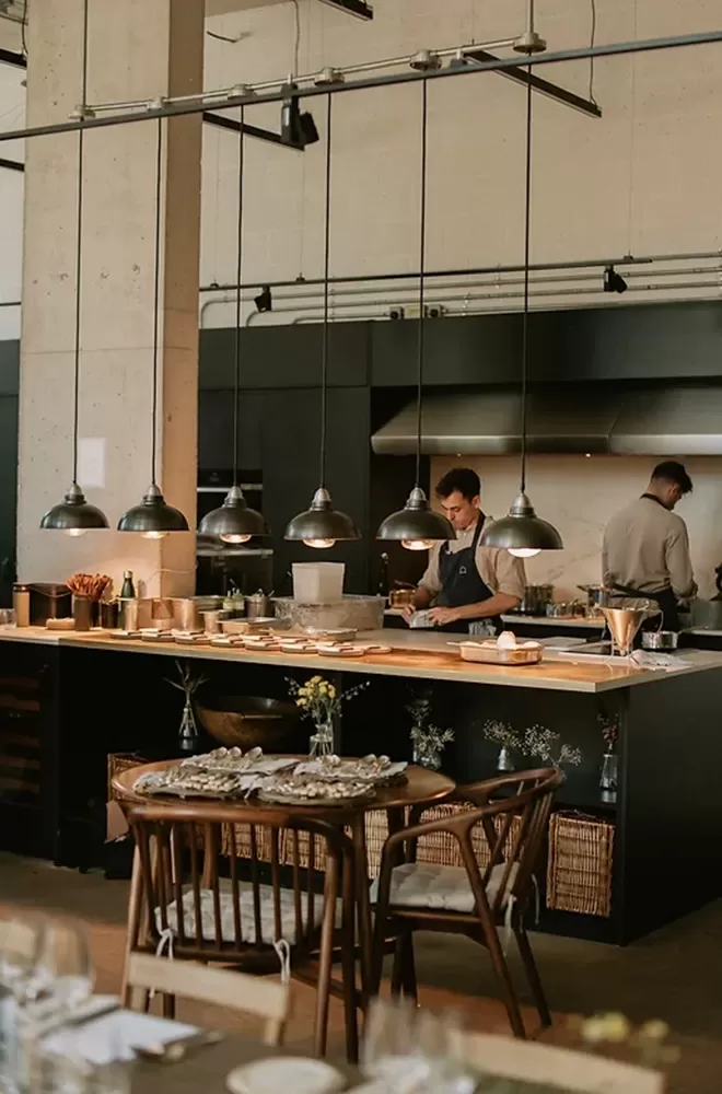 A modern kitchen with two chefs preparing food. The space has industrial-style pendant lights, wooden shelves, and neatly arranged kitchenware. A dining table with chairs and floral arrangements is in the foreground at The Water House Project, Bethnal Green.