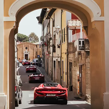 Line of red supercars travelling through arch and street in Italy during The Run To Monaco The Run To Monaco spectacular adventure through Italy