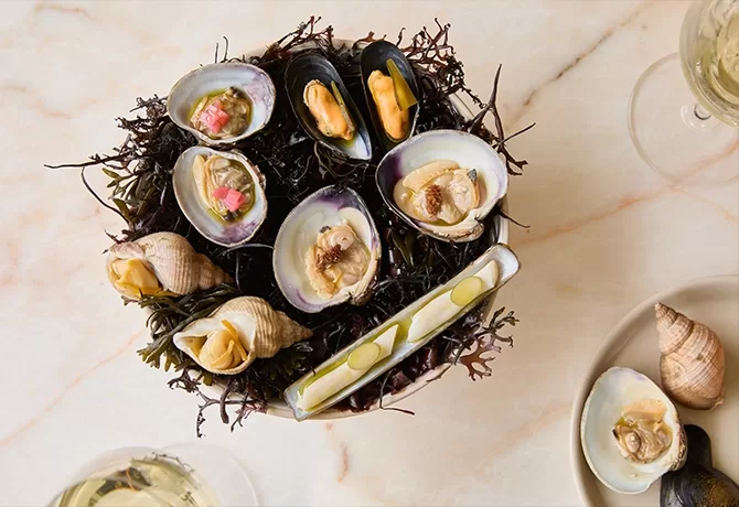 A plate of assorted fresh shellfish, including clams, mussels, whelks, and a razor clam, arranged on a bed of seaweed, viewed from above on a light marble surface. Glasses of white wine sit nearby.