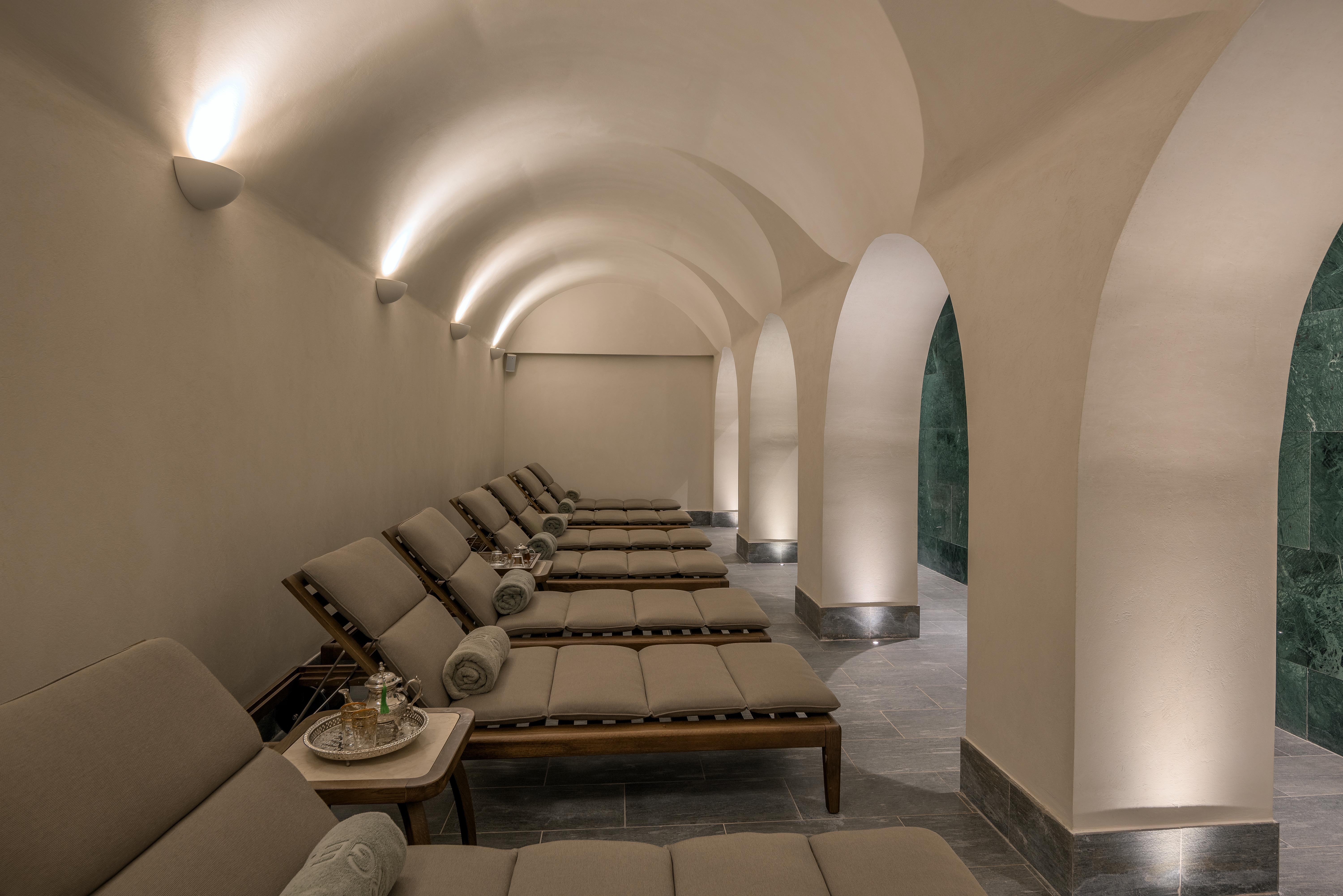 Inside spa with row of grey pillowed sunbeds with rolled up matching grey towels and bedside tables side a lit up white arch way.