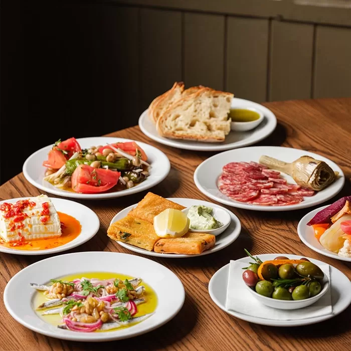 A variety of small Mediterranean dishes on white plates, including bread, cheese, olives, cured meats, tomatoes, vegetables, and dips, arranged on a wooden table.