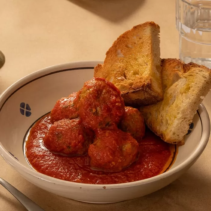 A bowl of meatballs in tomato sauce is served with several slices of toasted bread. A glass of water is visible in the background on a beige table.