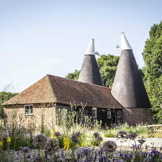 A traditional building with two conical roofs stands amid lush greenery. The structure has a brick facade with wooden elements, surrounded by wildflowers in the foreground under a clear sky at Tillingham Rye.