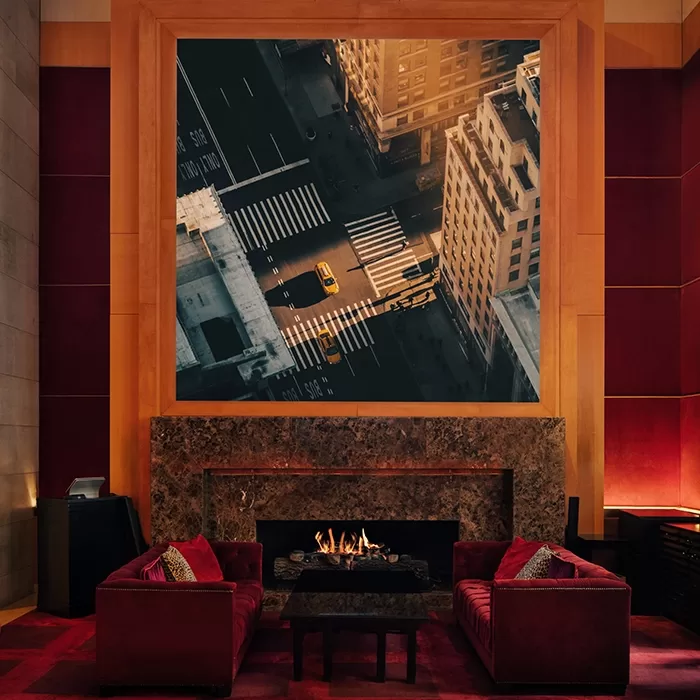 A cozy lounge with red velvet chairs and a lit fireplace, featuring a large framed photo above the mantel showing an aerial view of a city street with yellow taxis and tall buildings at sunset.