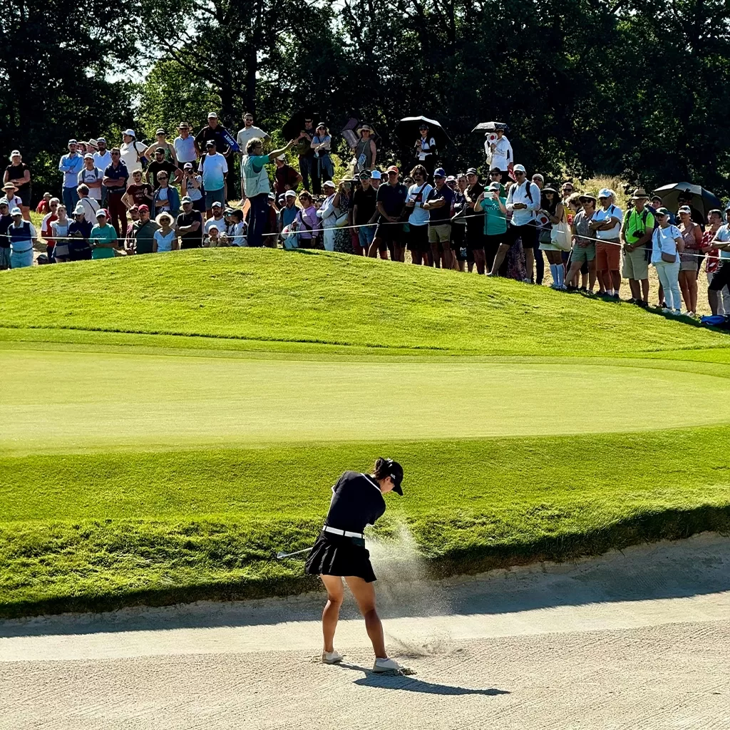 A golfer in a black outfit hits a shot from a sand bunker onto the green, with a large crowd of spectators watching from a grassy hill in the background. Sunlight casts strong shadows on the scene.
