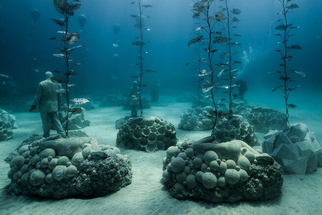 Under the sea sculpture park with multiple circular ball stacked sculptures spaced apart in piles with green sea plant sticking out of them and a man in a suit sculpture walking through them. Sculptures by by Jason Decaires Taylor