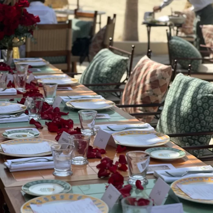 A long outdoor dining table is set with plates, menus, glasses, and napkins. Green and patterned cushions are on the chairs, and red rose petals are scattered along the center of the table. People are seen in the background.