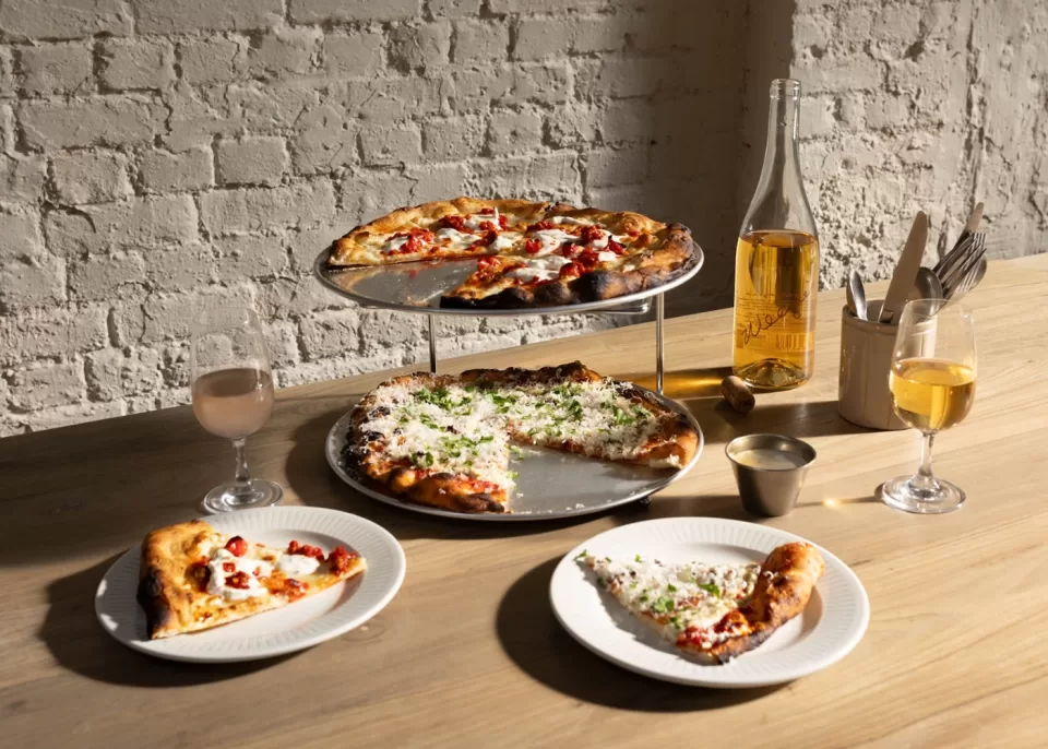 Two pizzas, one on a metal stand and one below it, are on a wooden table with two plated slices, two glasses of drinks, and a bottle. Sunlight casts shadows across the scene, highlighting the rustic brick wall in the background.