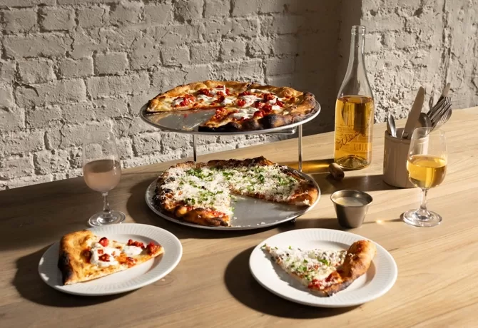 Two pizzas are displayed on a two-tier stand on a wooden table, with two slices served on separate plates. There are drinks in glasses, a wine bottle, a cup with utensils, and sunlight casts shadows on a white brick wall.