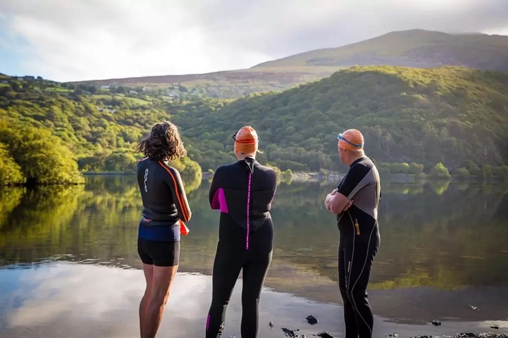 Women and two men dressed in wetsuits and swimming gear facing the view of Lake Windermere and the green hills surrounding it.