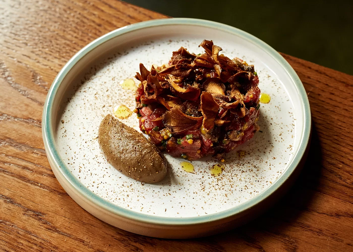 A round serving of beef tartare topped with crispy mushrooms sits on a white plate with a light brown rim, next to a dollop of creamy brown pâté, on a wooden table.