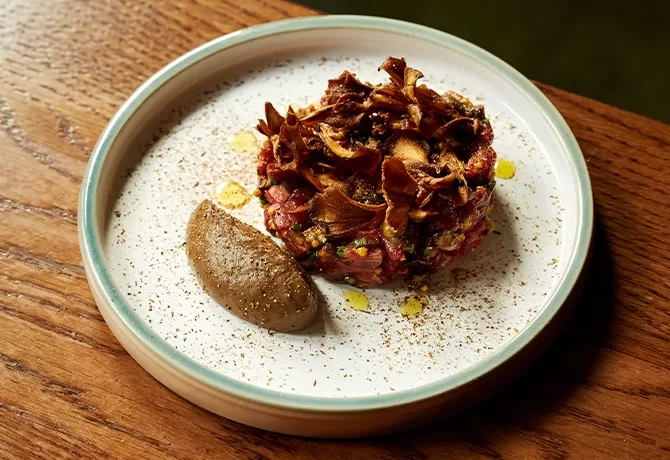 A round serving of steak tartare topped with crispy mushrooms sits on a white plate beside a dollop of creamy brown sauce, garnished with a drizzle of oil and a sprinkle of seasoning. The plate is on a wooden table.
