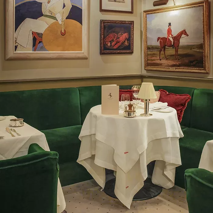 A cozy restaurant corner with green velvet seating, a round table set with a white tablecloth, elegant place settings, and framed artwork on the walls, including equestrian and abstract paintings.