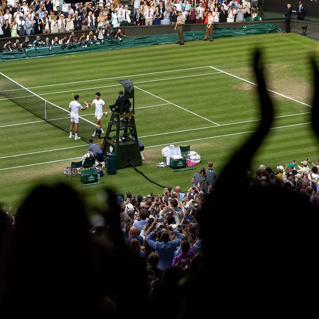 Two tennis players shake hands at the net after a match on a grass court, as a large, cheering crowd applauds from the stands, some with raised hands in the foreground.