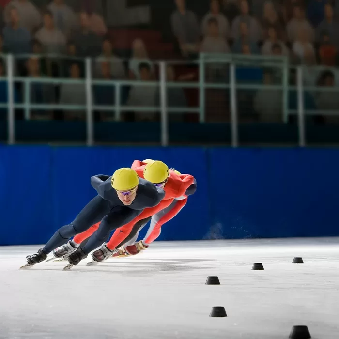 Two speed skaters wearing yellow helmets and racing suits compete closely on an indoor ice rink, leaning into a curve with blurred spectators visible in the background.