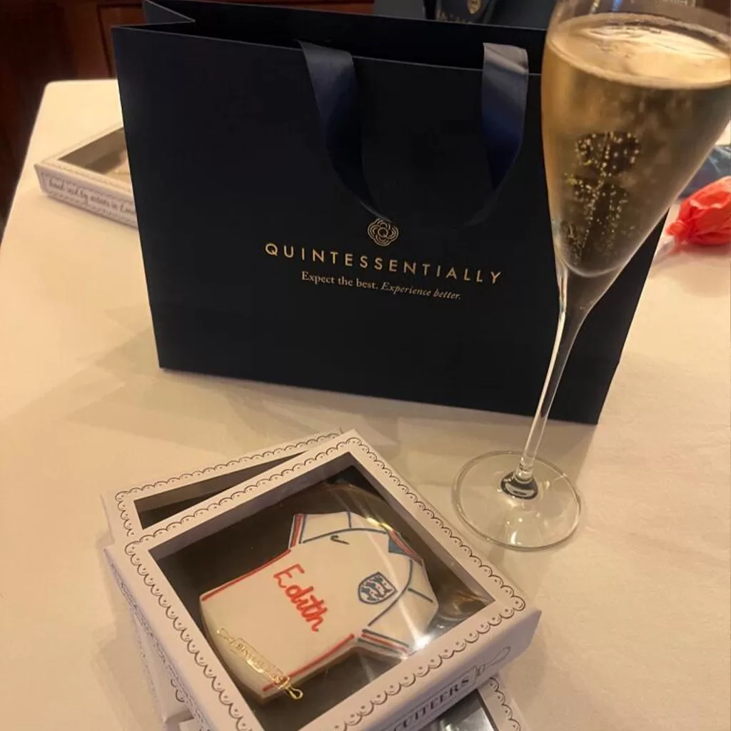 A navy Quintessentially gift bag, a glass of champagne, and a boxed shirt-shaped cookie with Edith written on it are displayed on a white tablecloth.