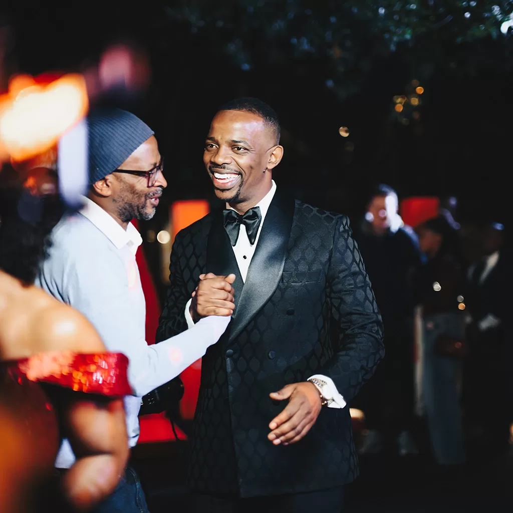 Two men greet each other warmly at a formal event. One man wears a patterned black tuxedo, while the other wears glasses, a beanie, and a light sweater. People and lights are blurred in the background.