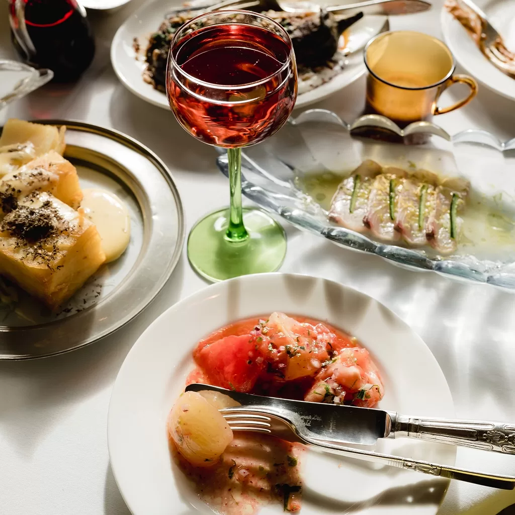 A table set with plates of gourmet food, including sliced tomatoes, a glass of red wine, a gold-rimmed cup, a plate with fish slices, and a dessert with sauce, all arranged under natural light.