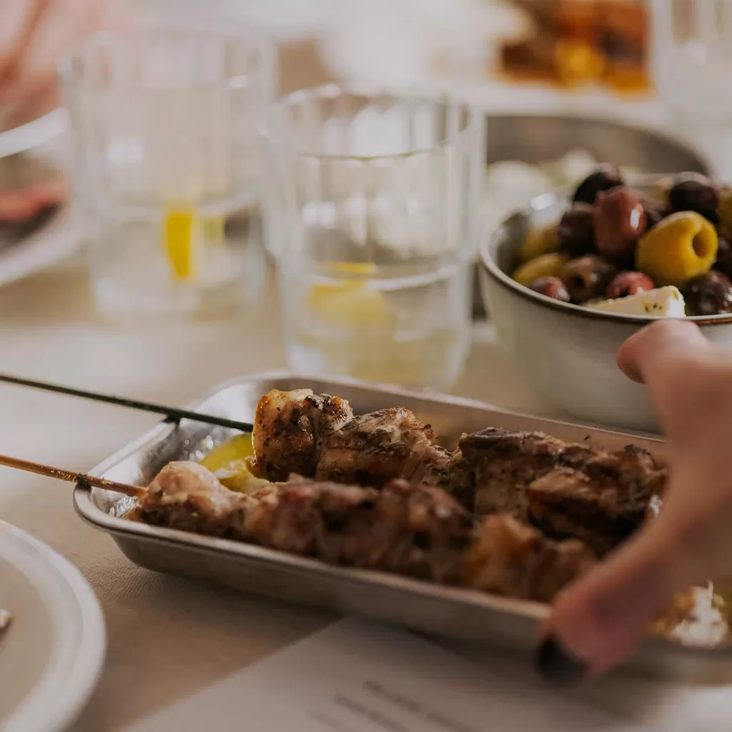 A close-up of skewered grilled meat on a metal tray, with a hand reaching for it. In the background, there are glasses of water with lemon and a bowl of mixed olives on a light-colored table.