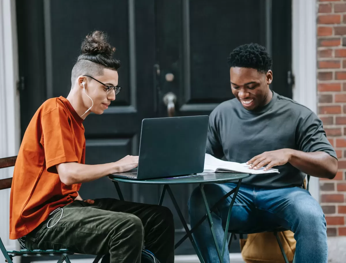 Student and tutor in casual clothing sat on an outdoor table in front of a building with a laptop and paper