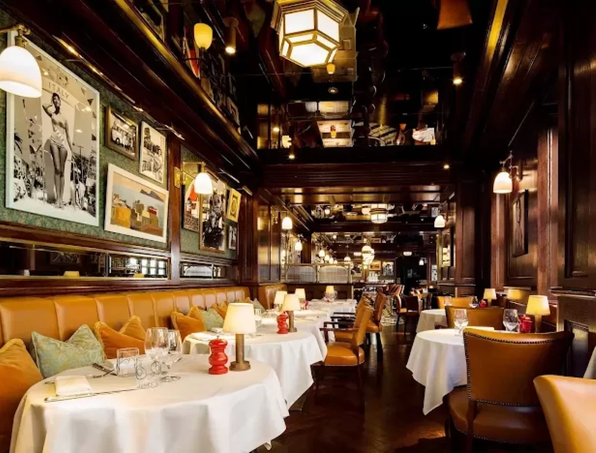A cozy, elegant restaurant with brown leather chairs, round tables with white tablecloths, warm lighting, and framed photos on dark wood-paneled walls. The atmosphere is inviting and upscale.