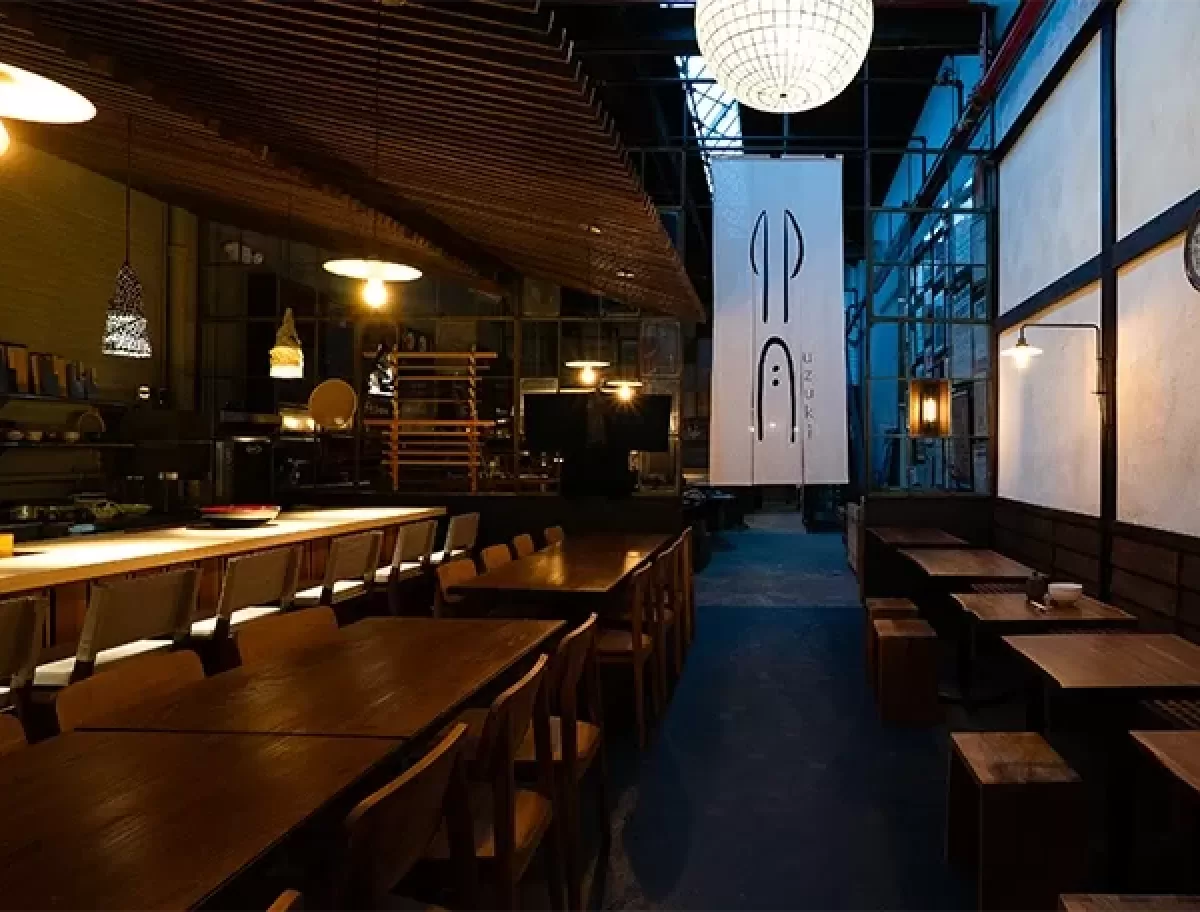 A dimly lit restaurant with wooden tables and chairs. A large, circular hanging light illuminates the space. A white banner with abstract symbols hangs near the back, and theres ambient, warm lighting throughout the interior.