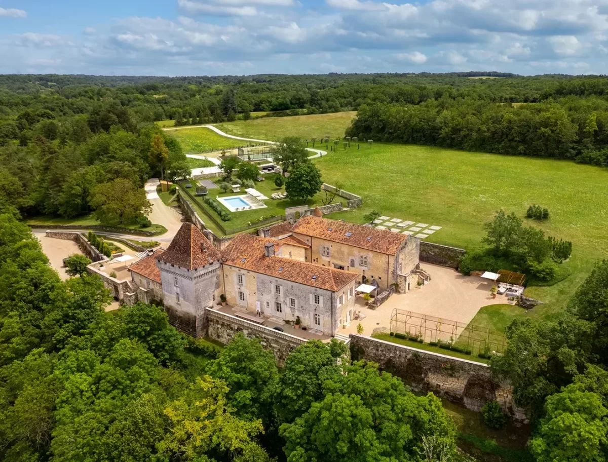 Aerial view of a historic chateau surrounded by lush greenery in Périgord, France. The building has a red-tiled roof and is bordered by stone walls. A swimming pool and landscaped gardens are visible. A winding road leads to the entrance. Rolling fields and trees extend beyond.