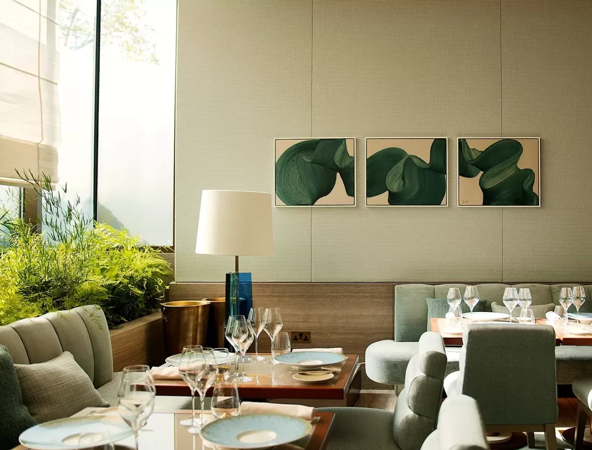 A modern dining room with green cushioned chairs and wooden tables set with glassware and blue-rimmed plates. A trio of abstract green artworks hang on the wall, and a large window lets in natural light, illuminating a potted plant nearby.