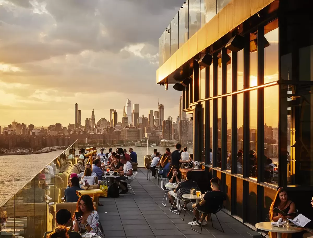 People dining and socializing on a rooftop terrace at sunset, with glass railings overlooking a city skyline full of tall buildings and skyscrapers under a partly cloudy sky.