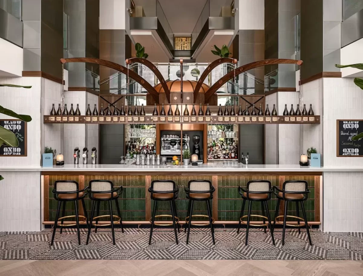 Modern bar interior with a long counter lined by six chic bar stools. Shelves stocked with bottles are displayed above, flanked by two chalkboards. The flooring has a geometric pattern, and large plants decorate the space.