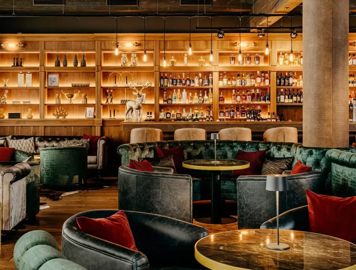A cozy lounge with leather chairs and plush sofas surrounds round tables. A well-lit wooden bar is lined with a variety of bottles, creating a warm and inviting atmosphere. Soft lighting enhances the ambiance.