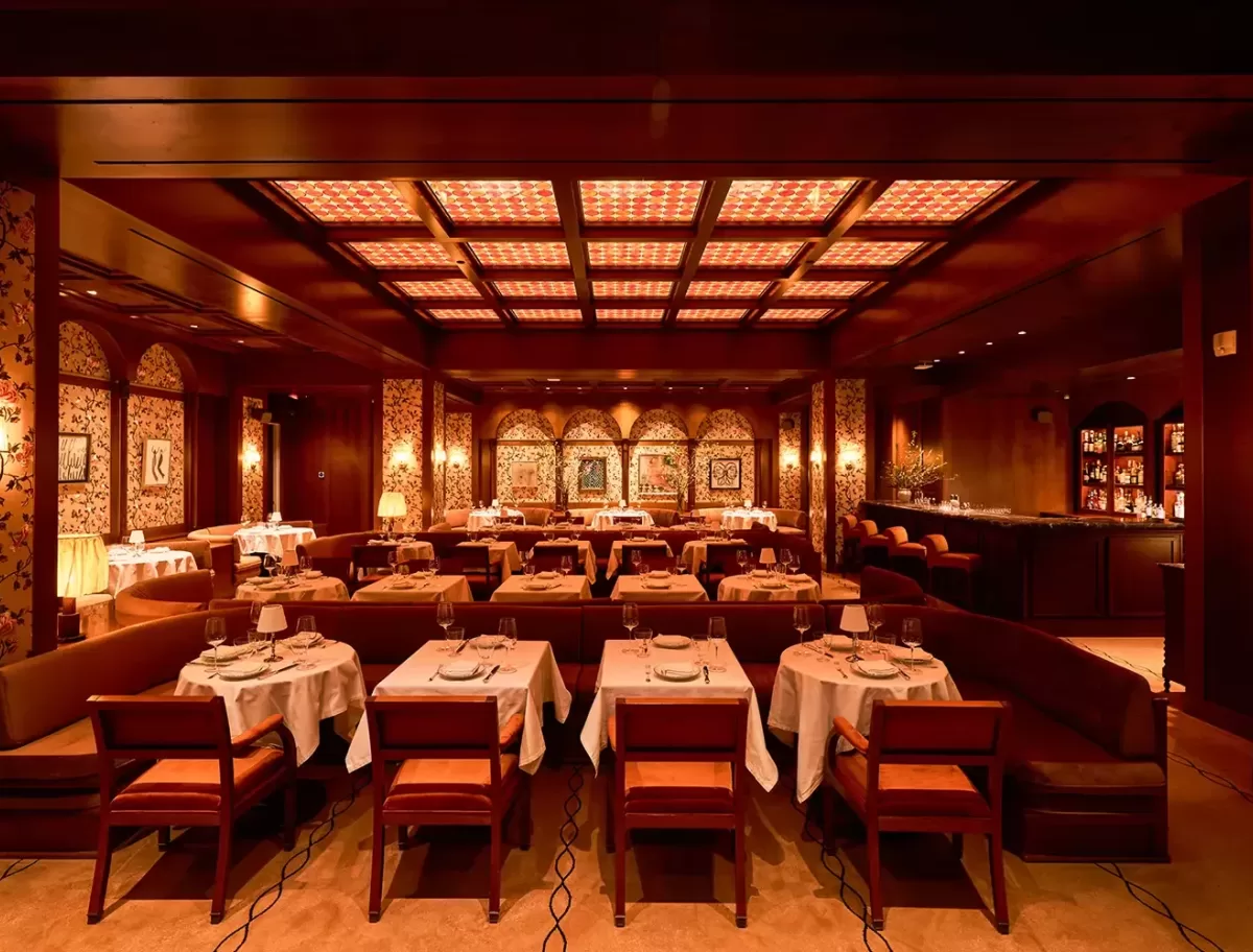 Elegant restaurant interior with warm lighting, featuring rows of tables set with white tablecloths and wooden chairs. The decor includes patterned wallpaper, framed artwork, and an inviting bar area in the background.
