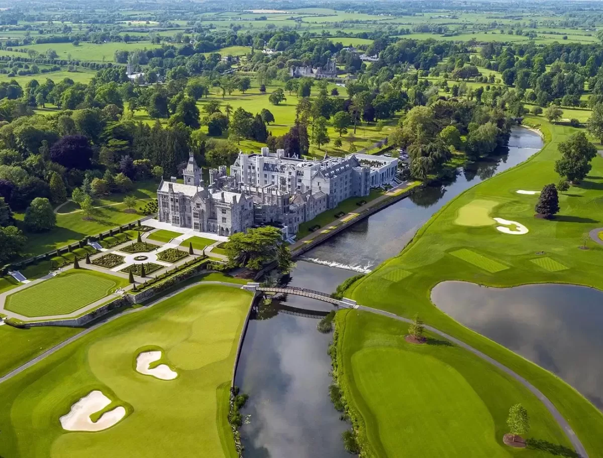 Aerial view of a large, historic castle-like mansion surrounded by lush green gardens, a moat, and expansive golf course with sand bunkers. The landscape is dotted with trees and stretches into the distance under a clear sky.
