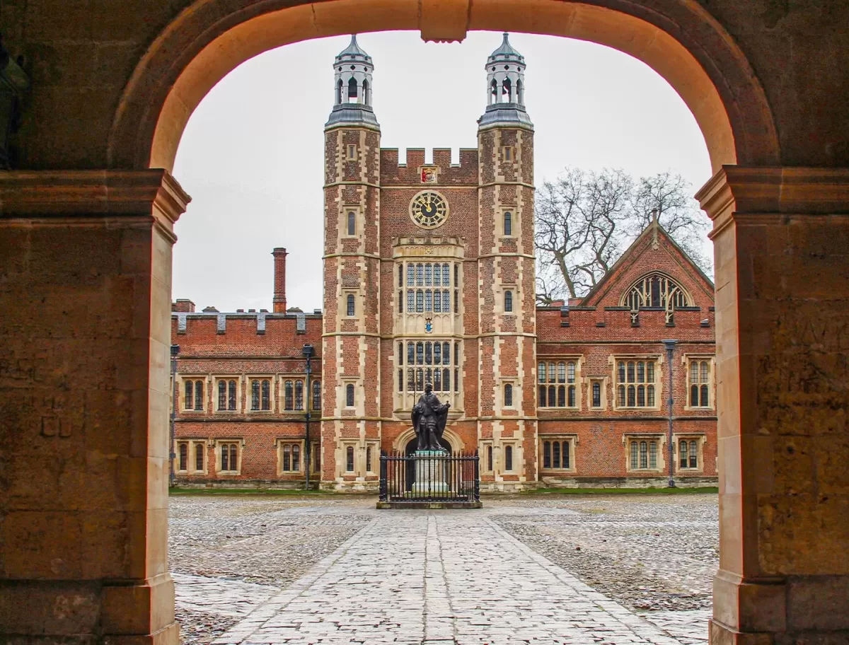 View through an archway of a historic brick building with a clock tower, central statue, cobblestone courtyard, and gothic architectural details on a cloudy day.