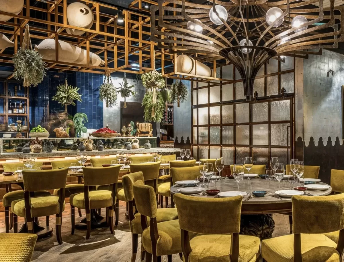 A warmly lit restaurant features tables set for dining with yellow upholstered chairs. Decorative plants hang from the ceiling. A wooden lattice design and a large artistic light fixture add to the cozy ambiance.