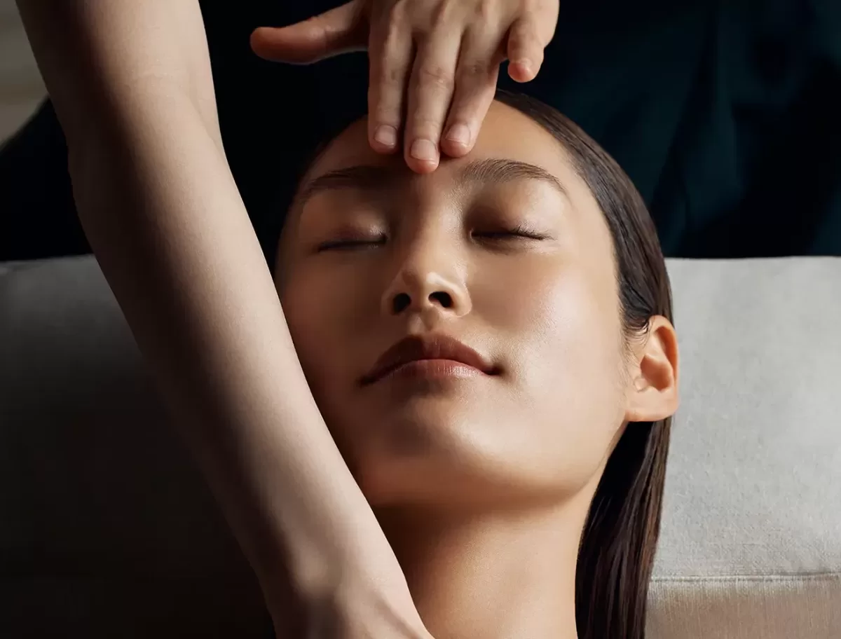 A person with closed eyes receiving a facial massage. Two hands gently press on the persons forehead and cheek. The setting is calm and relaxing, suggesting a spa or wellness environment at La Mer at the Ritz Carlton Hong Kong.