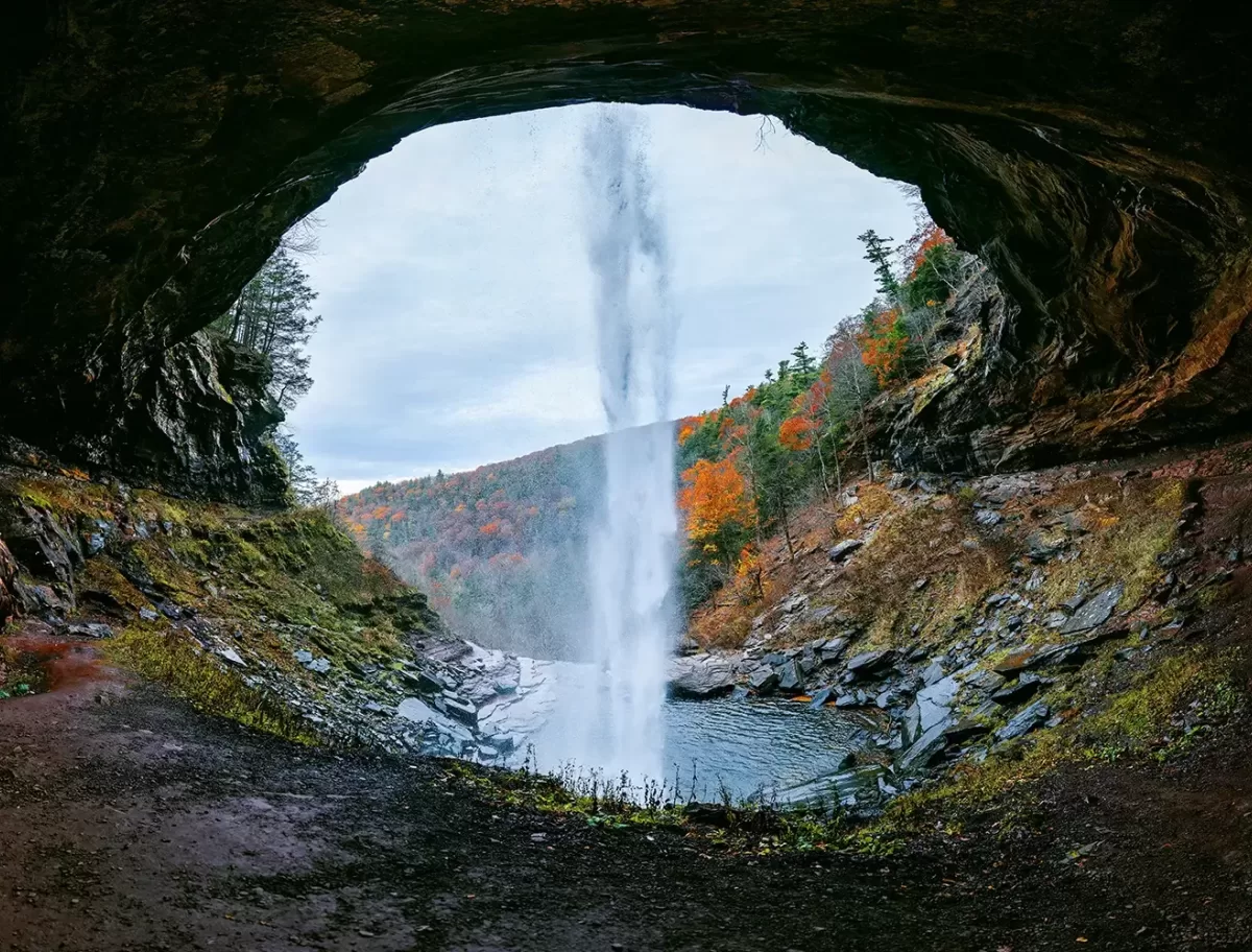 View from behind a waterfall cascading from a cave opening, with a serene pool below. The surrounding landscape is lush with autumn foliage in hues of orange and red, under an overcast sky.
