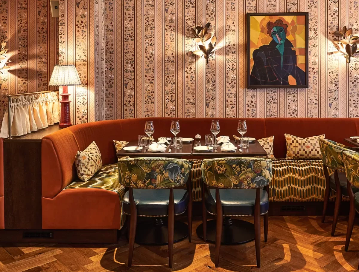 Cosy restaurant interior featuring a curved orange booth with patterned pillows, surrounded by green, floral-upholstered chairs, a patterned wallpaper, and a colorful abstract painting. Tables are set with glasses and napkins.