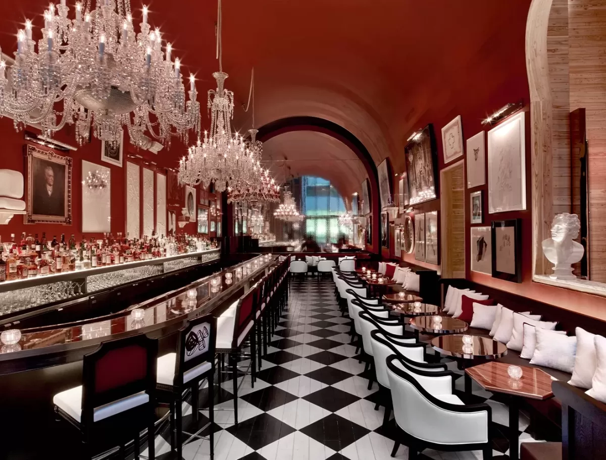 Luxurious restaurant interior with elegant chandeliers, a long bar on the left, and rows of tables with white chairs on the right. The room features a high ceiling and red walls adorned with framed artwork and mirrors. Black and white checkered floor tiles.