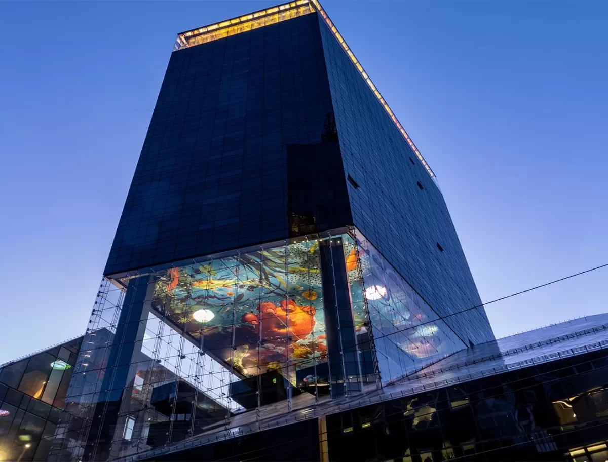 A modern, tall building with a reflective glass facade is shown against a clear blue sky. A vibrant digital display underneath a protruding section features colorful abstract art, visible from the street below.