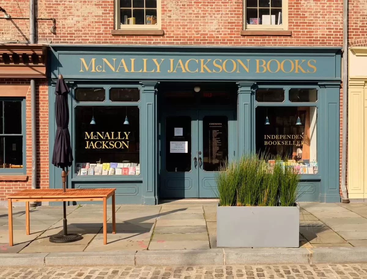 Exterior of McNally Jackson Books in NYC with a green facade and gold lettering. The storefront features large windows displaying books, a small table, a closed umbrella, and a planter with tall grasses on the cobblestone sidewalk.