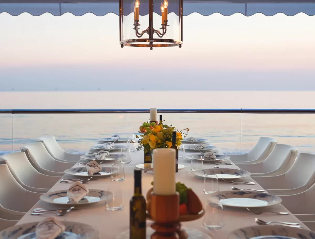 A long dining table set for a meal with white chairs, plates, glasses, and floral centerpieces sits on a terrace overlooking the calm sea at sunset. A chandelier hangs above the table.