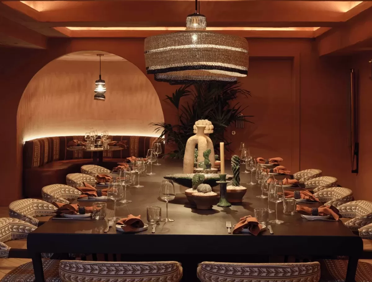 A dimly lit, elegantly set dining room with a long table surrounded by patterned chairs. A large decorative light fixture hangs above, and the table is adorned with glassware, orange napkins, and intricate centerpieces. The walls feature warm brown tones at Ixchel.