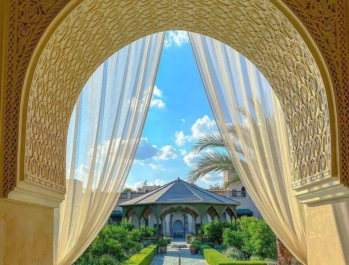 View through an ornate arched doorway with sheer curtains revealing a lush garden, palm trees, and a pavilion under a bright blue sky with scattered clouds. Intricate patterns decorate the arch.