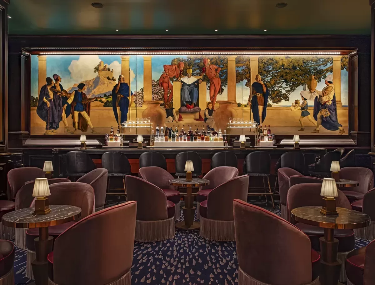 A dimly lit bar with plush velvet chairs and small tables equipped with lamps. A large, colorful mural depicting a historical scene is behind the well-stocked bar counter, adding an artistic touch to the sophisticated setting.