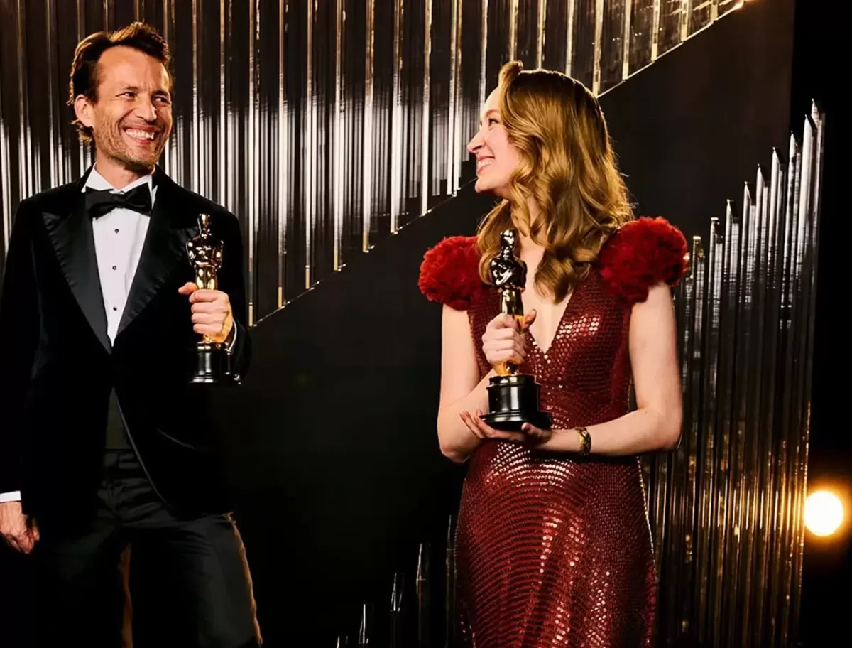 A man and a woman in formal attire hold trophies and smile at each other. The man wears a black tuxedo, and the woman is in a red sparkling dress with feathered shoulders. They stand against a stylish, dark background with vertical lines.